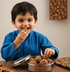 A young boy in a blue shirt smiles while eating a nut and fruit energy ball from an open round metal tin filled with similar balls, set on a wooden table.