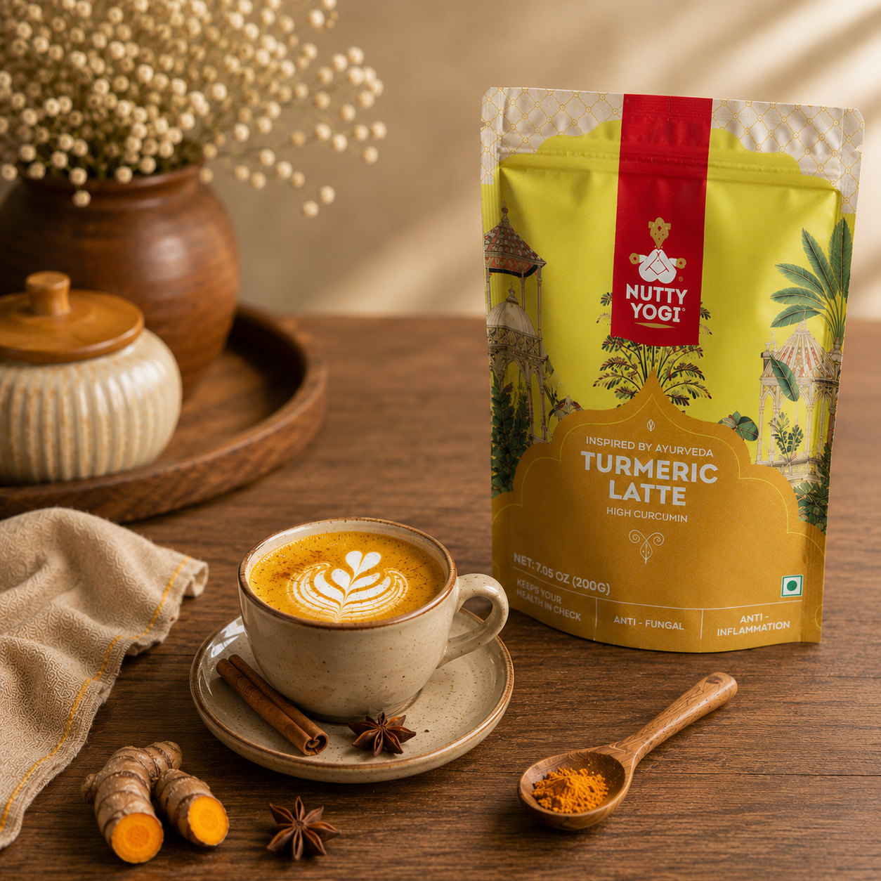 A stand-up pouch of Nutty Yogi Turmeric Latte tea mix stands on a wooden table beside a cup of latte with leaf latte art, cinnamon sticks, star anise, turmeric roots, and a small spoon of orange powder. The pouch is yellow and gold with a red vertical label and illustrated tropical scenery.