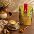 A stand-up pouch of Nutty Yogi Turmeric Latte tea mix stands on a wooden table beside a cup of latte with leaf latte art, cinnamon sticks, star anise, turmeric roots, and a small spoon of orange powder. The pouch is yellow and gold with a red vertical label and illustrated tropical scenery.