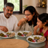 A family of three sharing a meal together at a glass table, with plates of rice, curry, pickle, and flatbread. The father on the left wears a white kurta, the mother in the center wears glasses and a light beige top, and the young girl on the right wears a red and green traditional outfit.