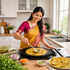 A woman in a yellow and magenta saree cooks a flatbread on a black kadhai pan in a bright, modern kitchen. Small bowls of ingredients and a plate of finished flatbreads are arranged on the wooden countertop.