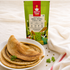 A package of Nutty Yogi Green Moong Cheela Mix stands behind a white plate stacked with golden-brown cheelas on a light fabric surface, with a folded white cloth and a few green herb leaves nearby.