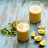 Two clear glasses filled with a yellow-green smoothie or juice on a distressed blue wooden surface, surrounded by lime wedges and cilantro.