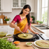 A woman in a yellow and magenta saree is cooking a thin green pancake (dosa) on a stovetop in a bright, modern kitchen. Bowls of ingredients, fresh cilantro, and a plate of finished dosas are arranged on the wooden countertop.