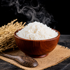 A wooden bowl filled with steaming white rice, set on a woven burlap mat with a wooden spoon and dried rice stalks against a black background.