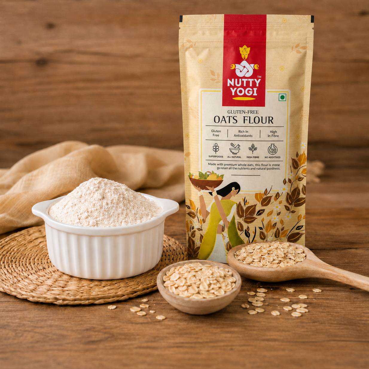 A stand-up pouch of Nutty Yogi Gluten Free Oats Flour stands on a wooden surface beside a white bowl filled with flour, wooden spoons with oats, and a woven mat. The packaging is beige with a red brand panel and leaf illustrations.