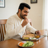 A man in a cream-colored kurta sits at a wooden dining table, smiling as he eats from a stainless steel plate containing a flatbread, sambar, chutney, and curry. A plate of sliced cucumbers and carrots is in the foreground, with a glass of water on the side.
