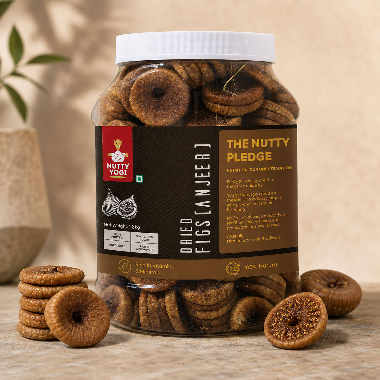 A large clear plastic jar of Nutty Yogi dried figs (anjeer) with a white lid and a dark brown label, displayed on a stone surface with loose dried figs around it. The background is a warm beige wall with soft shadows.