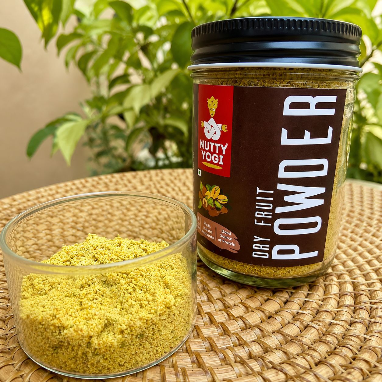 A glass container with dry fruit powder, next to a plastic jar labeled 'Nutty Yogi Nutri Dry Fruit Powder' on a woven mat.