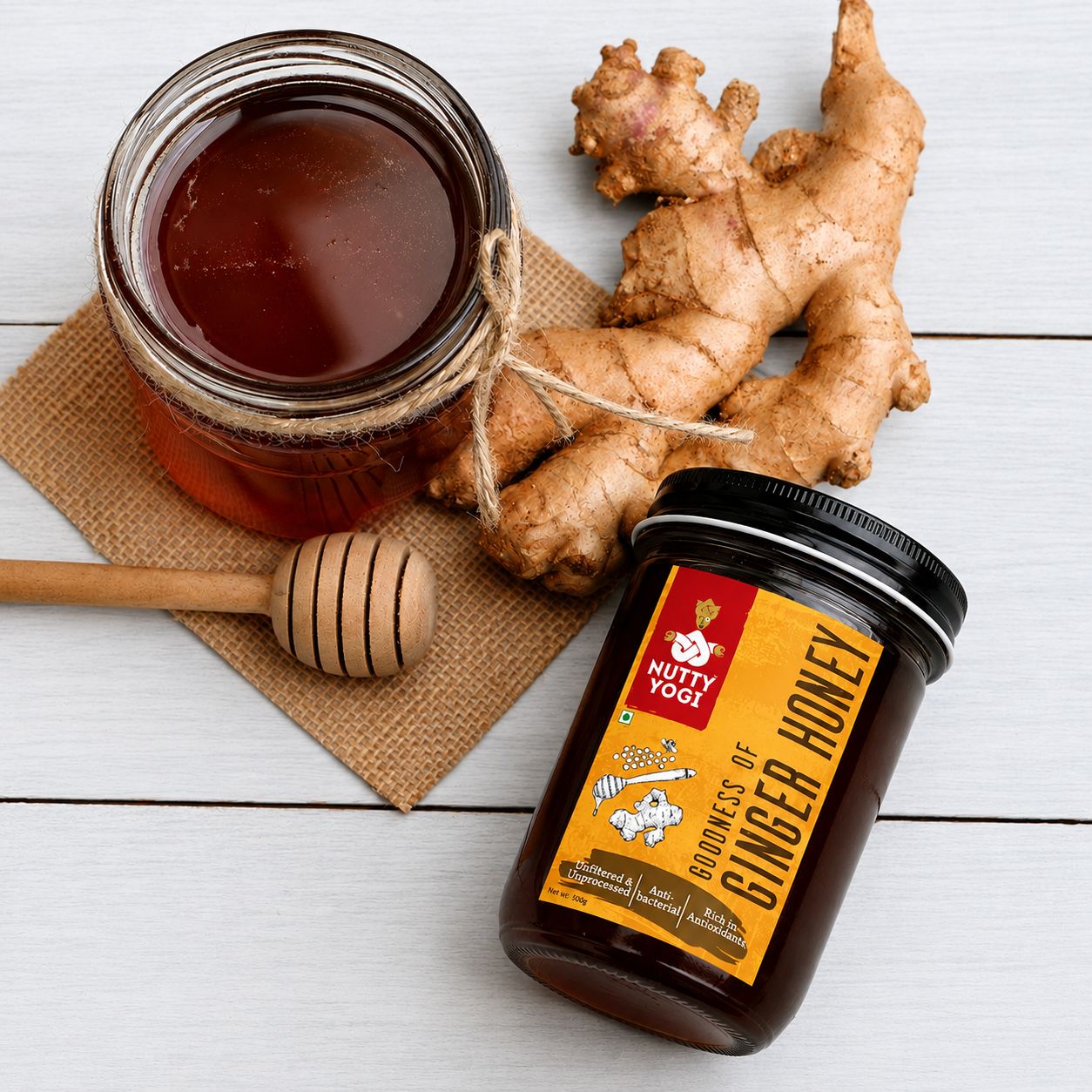 Nutty Yogi Goodness of Ginger Honey displayed alongside a glass jar of honey, fresh ginger roots, and a wooden honey dipper on a white wooden surface, highlighting a natural blend of ginger and honey rich in antioxidants.
