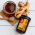Nutty Yogi Goodness of Ginger Honey displayed alongside a glass jar of honey, fresh ginger roots, and a wooden honey dipper on a white wooden surface, highlighting a natural blend of ginger and honey rich in antioxidants.