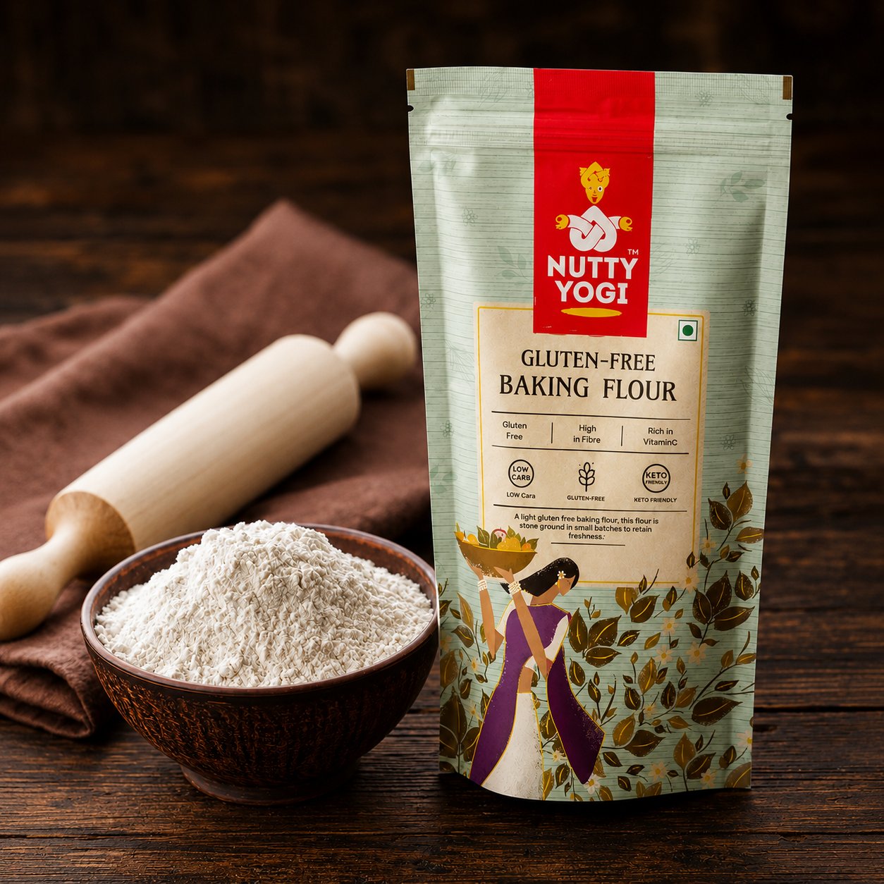 A stand-up pouch of Nutty Yogi Gluten-Free Baking Flour stands on a dark wooden surface next to a small bowl filled with white flour and a wooden rolling pin on a brown cloth.