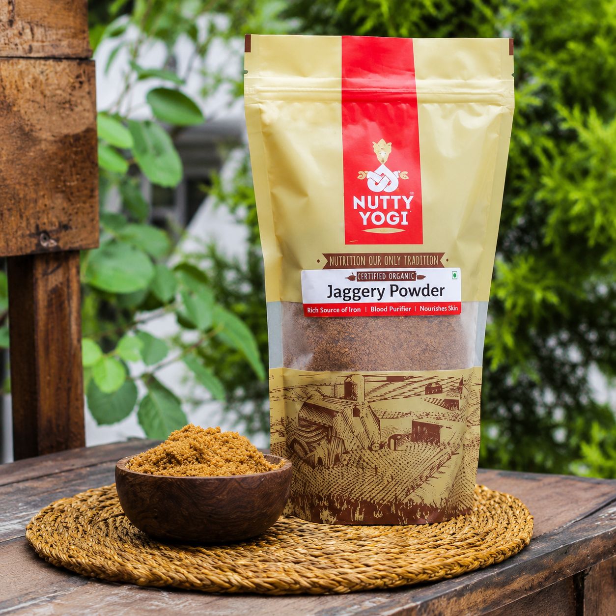 Nutty Yogi Jaggery Powder in a stand-up pouch displayed beside a bowl of natural jaggery powder on a woven mat with a garden background.