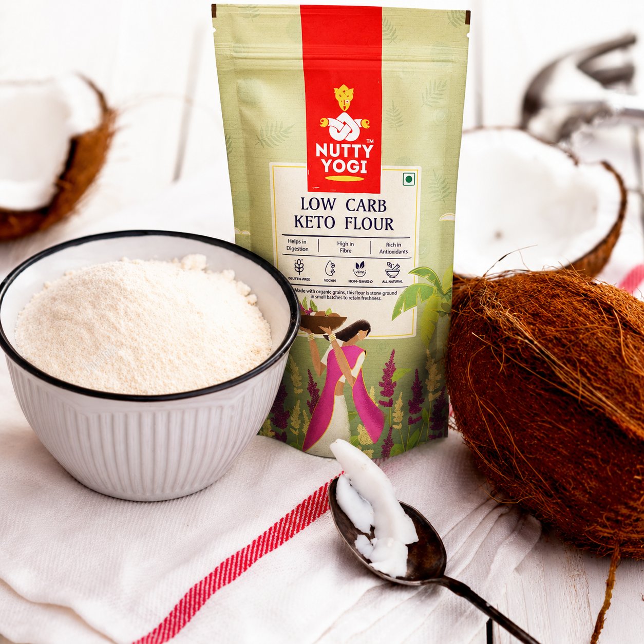 A pouch of Nutty Yogi Low Carb Keto Flour stands on a white cloth beside a bowl filled with white flour, a spoon with coconut, and whole/coconut halves, suggesting ingredients for keto nutrition.