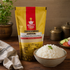 A stand-up pouch of Nutty Yogi Sona Masoori Rice (Unpolished) stands on a wooden table next to a bowl of cooked white rice, with a plant and brass containers in the background.