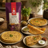 A package of Nutty Yogi Multigrain Missi Atta stands on a rustic wooden table surrounded by prepared flatbreads and dosas on plates, with small bowls of chutney and garnish. The background is softly blurred with indoor decor.