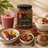 A jar of dried goji berry centered on a styled tabletop with bowls of yogurt, oatmeal, a smoothie, nuts, and a muffin. The jar label reads 'Dried GOJIERRY' and shows the brand logo at the top.