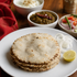 A stack of round toasted flatbreads on a white plate, served with sliced white onions and a lemon wedge, with bowls of chutney, pickle, and sliced red onions on a wooden table.