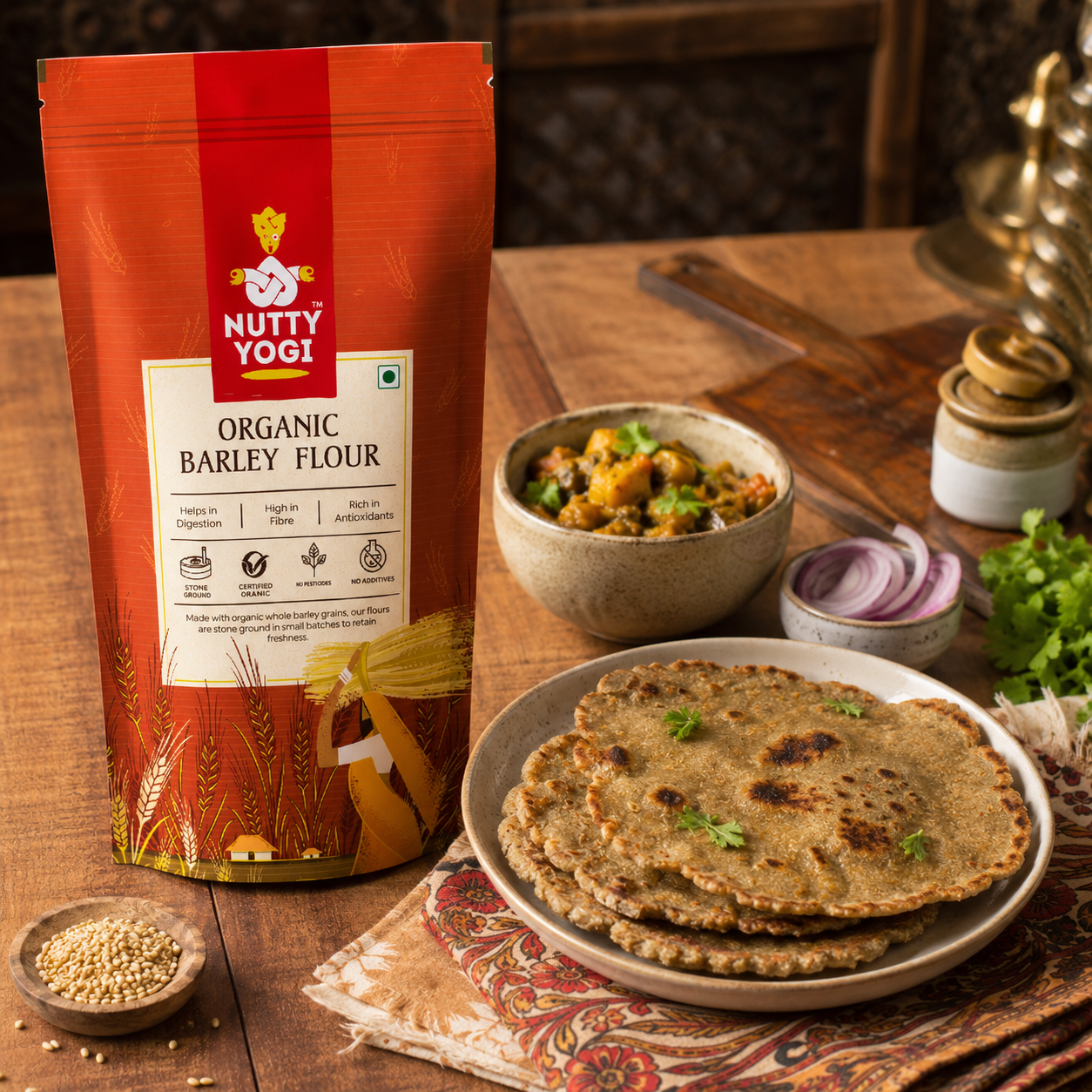 A bag of Nutty Yogi Organic Barley Flour stands on a wooden table beside a plate of flatbreads, a bowl of cooked dish, sliced red onion, cilantro, and small bowls of grains, creating a rustic kitchen scene.