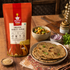 A bag of Nutty Yogi Organic Barley Flour stands on a wooden table beside a plate of flatbreads, a bowl of cooked dish, sliced red onion, cilantro, and small bowls of grains, creating a rustic kitchen scene.