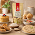 A bag of Nutty Yogi Gluten Free Oats Flour stands on a wooden table surrounded by prepared oatmeal dishes, including pancakes, muffins, flatbread, cookies, and a bowl of fruit yogurt, in a warm kitchen setting.